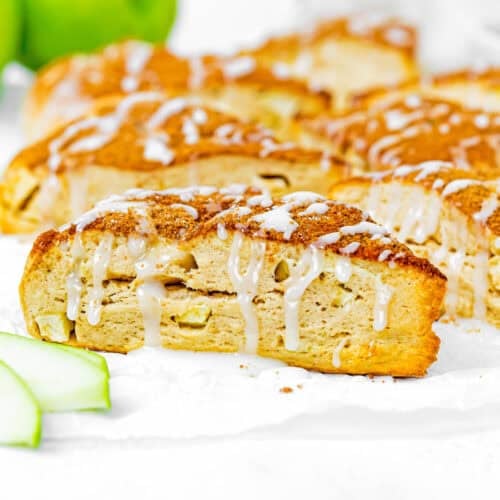 A close-up of Eggless Apple Cinnamon Scones, sliced and drizzled with white icing, with green apple slices and a whole green apple in the background.