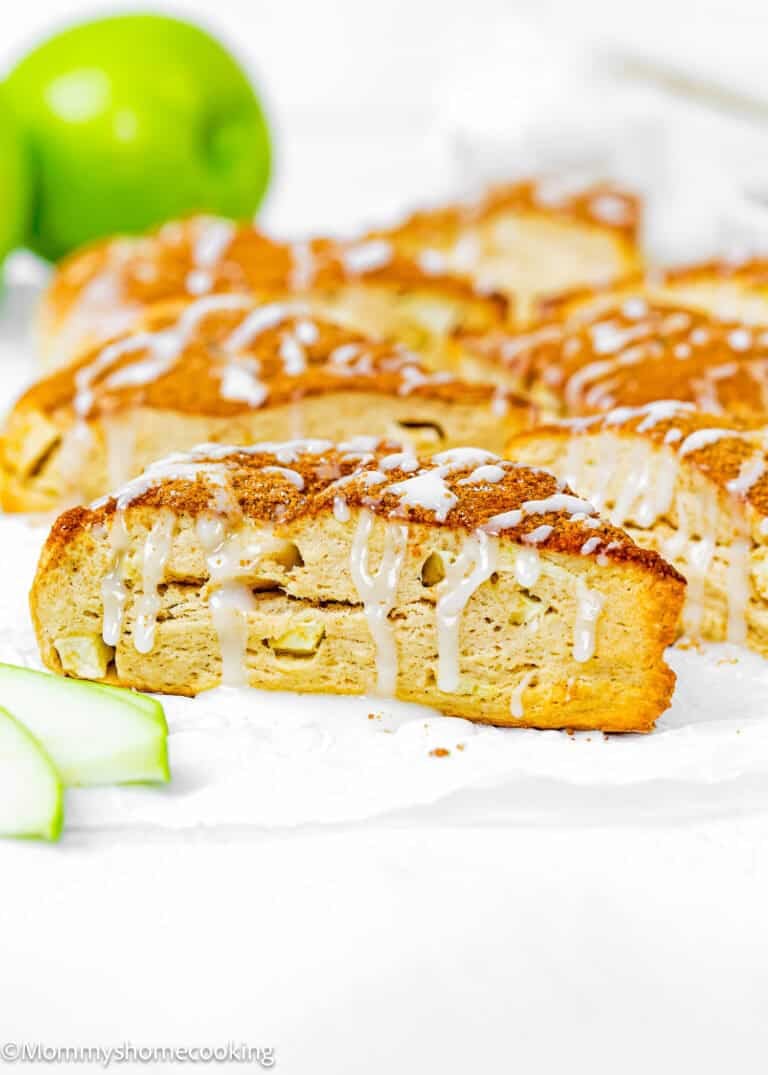 A close-up of Eggless Apple Cinnamon Scones, sliced and drizzled with white icing, with green apple slices and a whole green apple in the background.