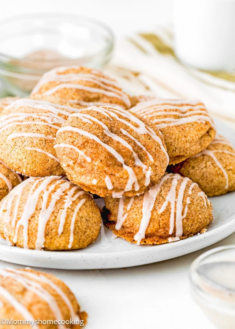 A plate of round, golden-brown cookies topped with white icing drizzle, arranged on a white surface with glass bowls and a glass of milk in the background.