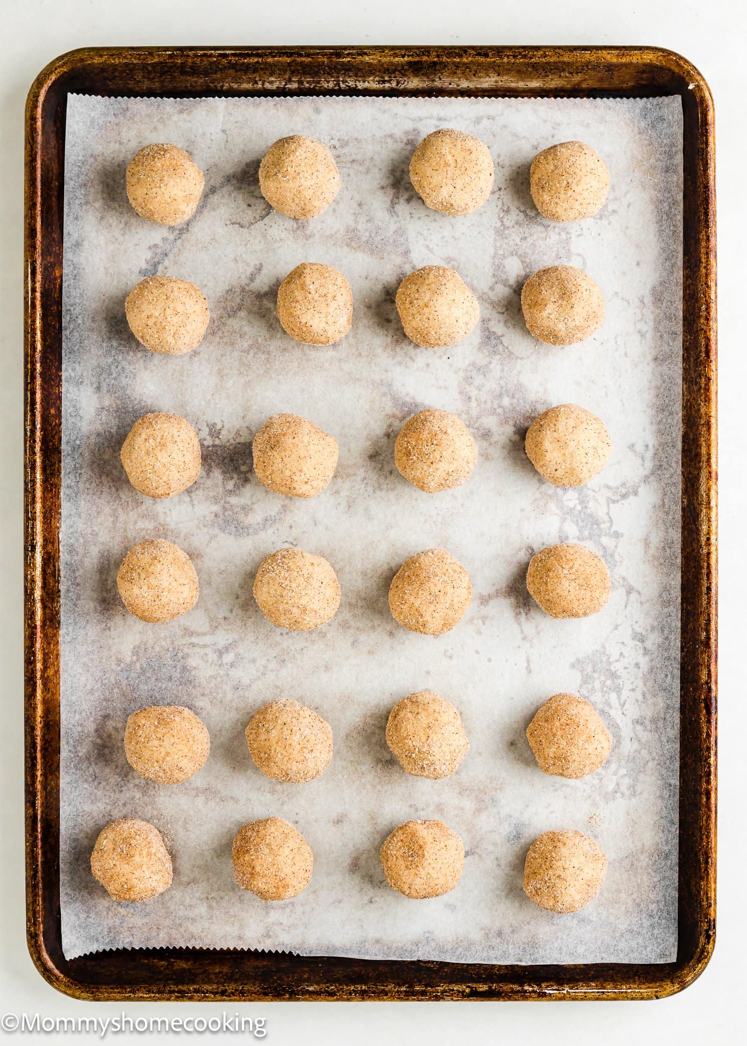 A baking sheet lined with parchment paper holds 20 evenly spaced balls of Eggless Apple Pie Snickerdoodle Cookies dough, each coated in cinnamon sugar.