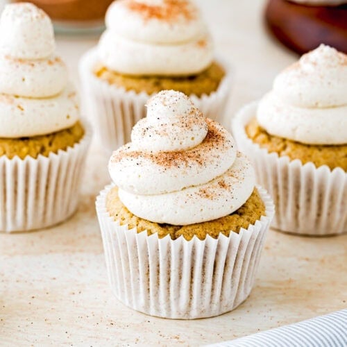 Four Eggless Banana Cupcakes with swirls of creamy frosting and a sprinkle of cinnamon on top are arranged on a light surface, with a striped napkin and a jar in the background.