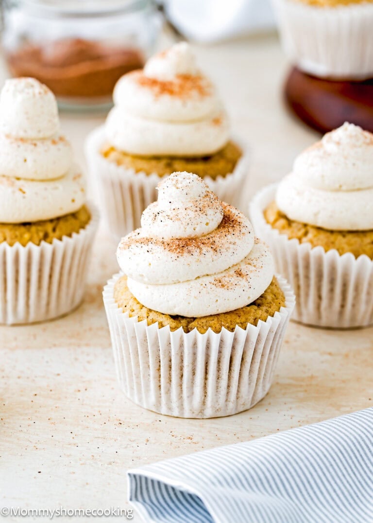 Four Eggless Banana Cupcakes with swirls of creamy frosting and a sprinkle of cinnamon on top are arranged on a light surface, with a striped napkin and a jar in the background.