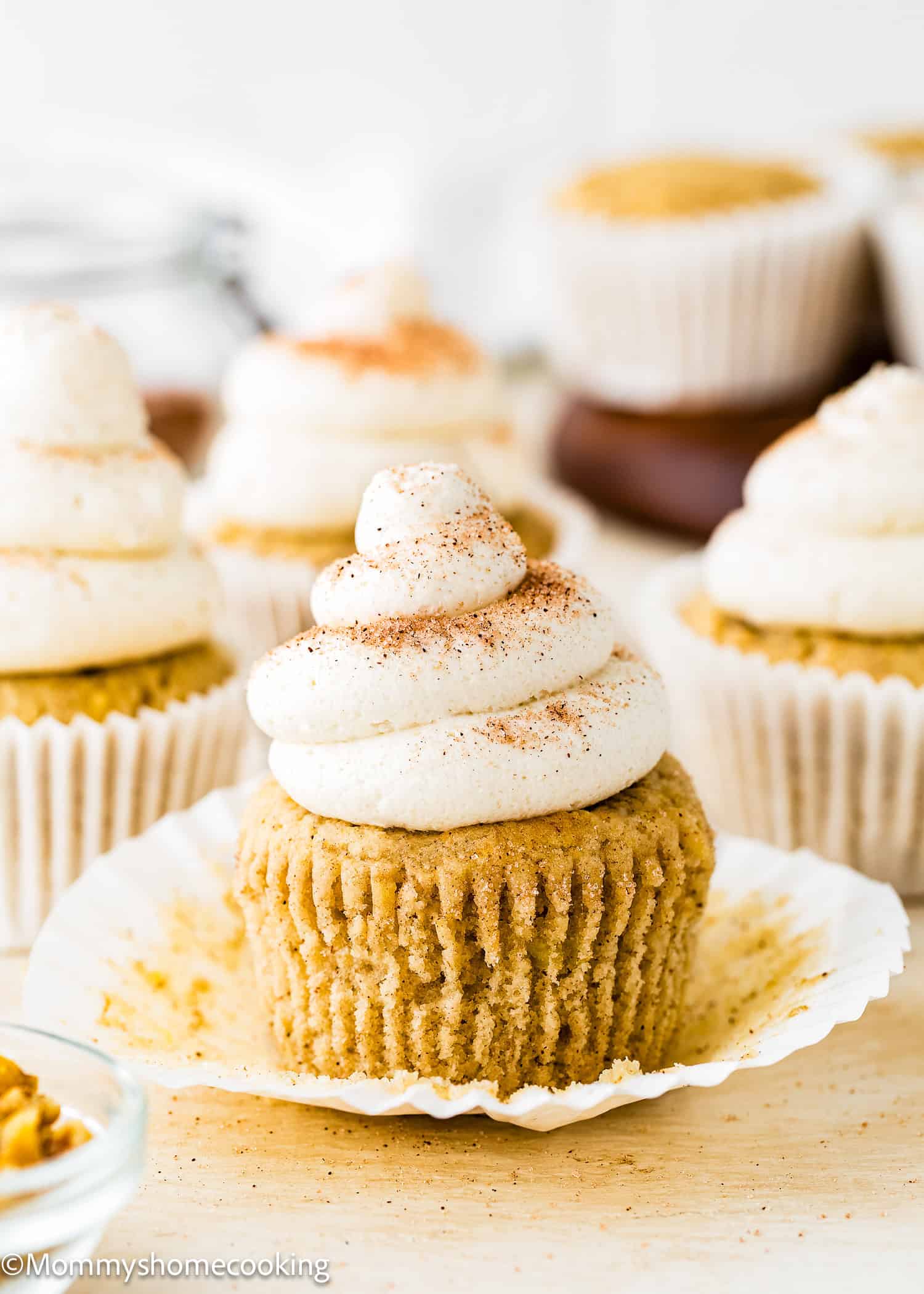 A egg-free banana cupcake with swirled white frosting and a sprinkle of cinnamon sits on an unwrapped paper liner, surrounded by more Eggless Banana Cupcakes decorated in a similar style.