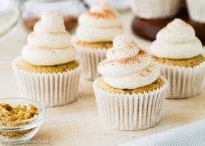 Four Eggless Banana Cupcakes with swirled white frosting and a sprinkle of cinnamon on top are arranged on a light surface. A small bowl of chopped nuts sits in the foreground.