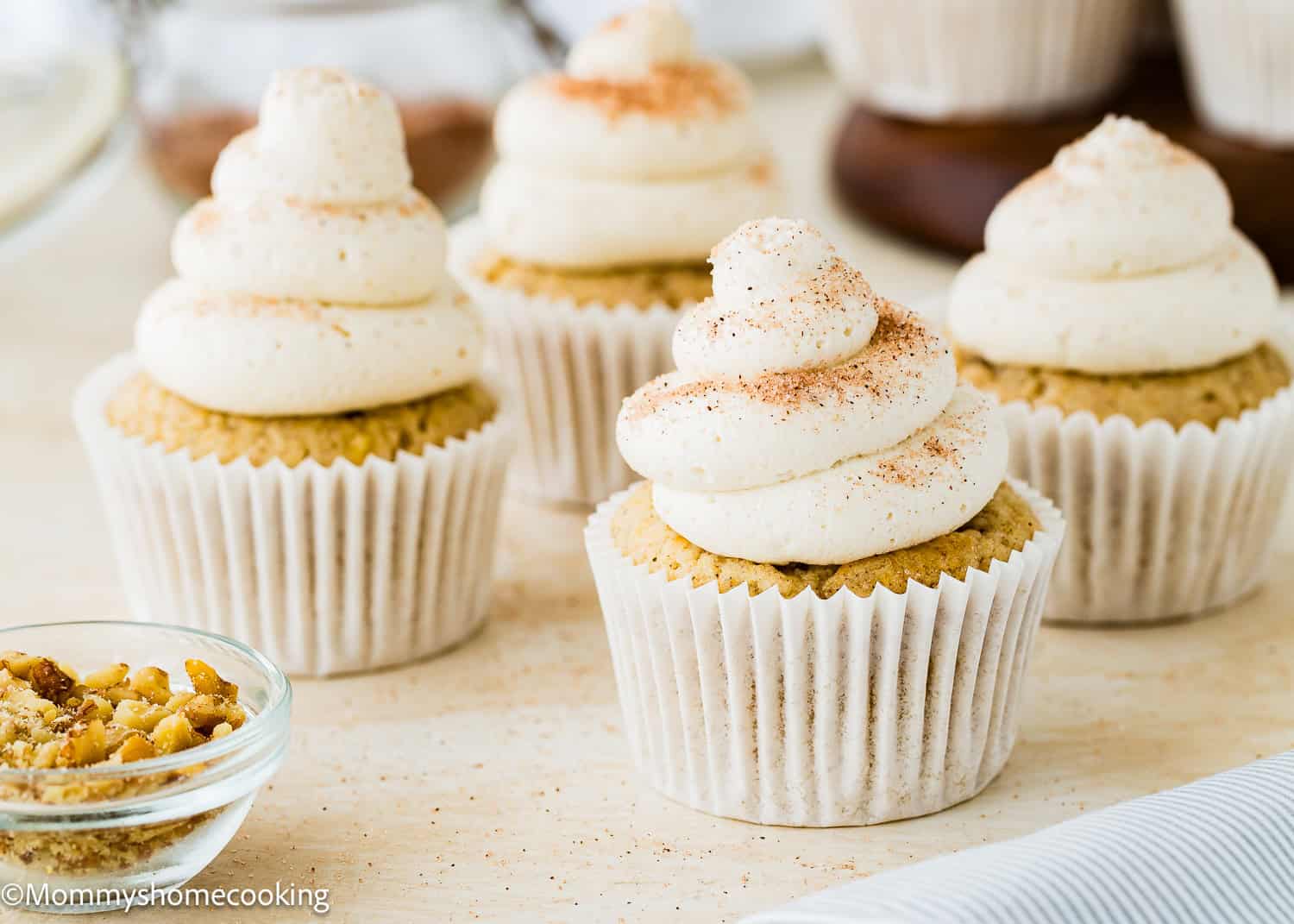 Four Eggless Banana Cupcakes with swirled white frosting and a sprinkle of cinnamon on top are arranged on a light surface. A small bowl of chopped nuts sits in the foreground.