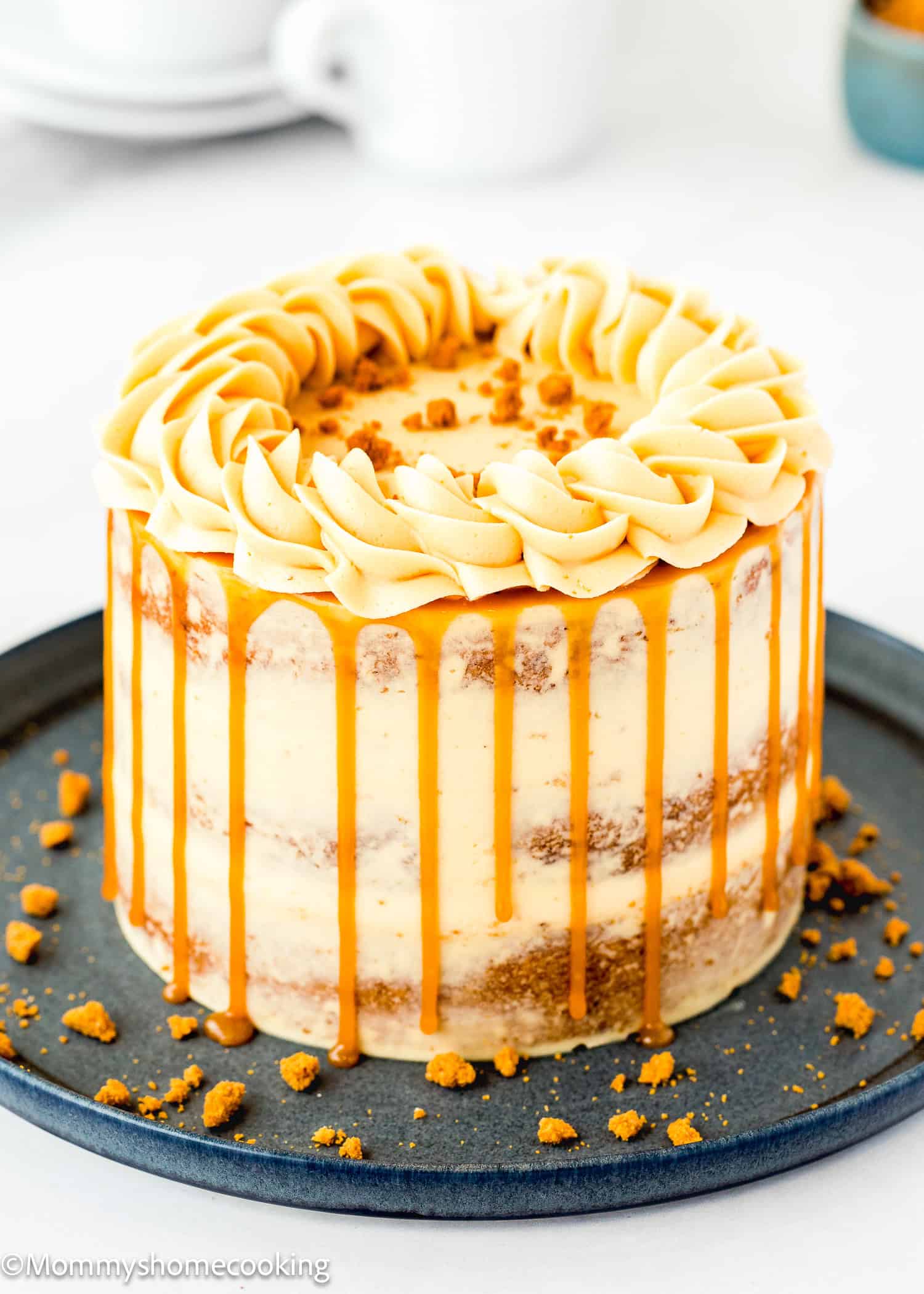 An Eggless Banana Layer Cake with light frosting and caramel drips sits on a dark plate, topped with piped swirls and cookie crumbs. Crumbled cookies are scattered around the base, adding an inviting touch.