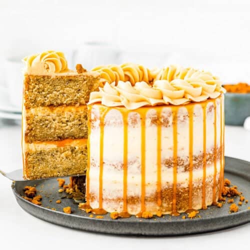 An Eggless Banana Layer Cake with caramel drizzle and piped frosting sits on a dark plate. A slice is being lifted, revealing moist banana layers and creamy filling inside. Blurred cups and a small bowl are in the background.
