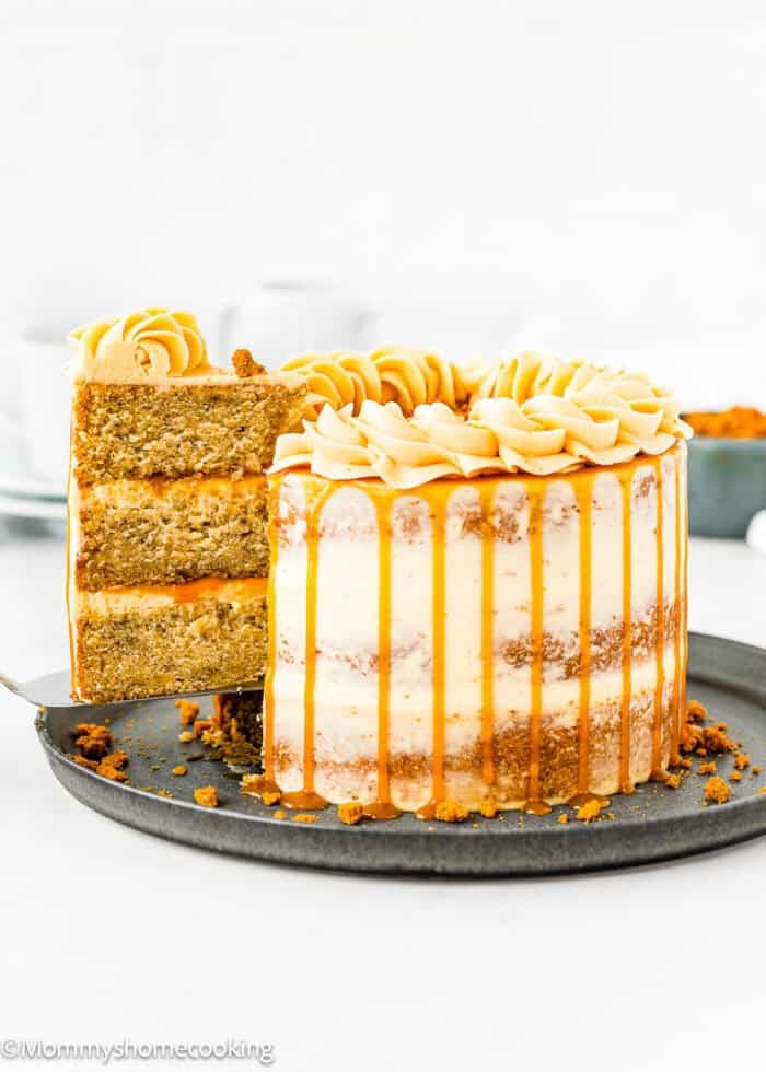 An Eggless Banana Layer Cake with caramel drizzle and piped frosting sits on a dark plate. A slice is being lifted, revealing moist banana layers and creamy filling inside. Blurred cups and a small bowl are in the background.