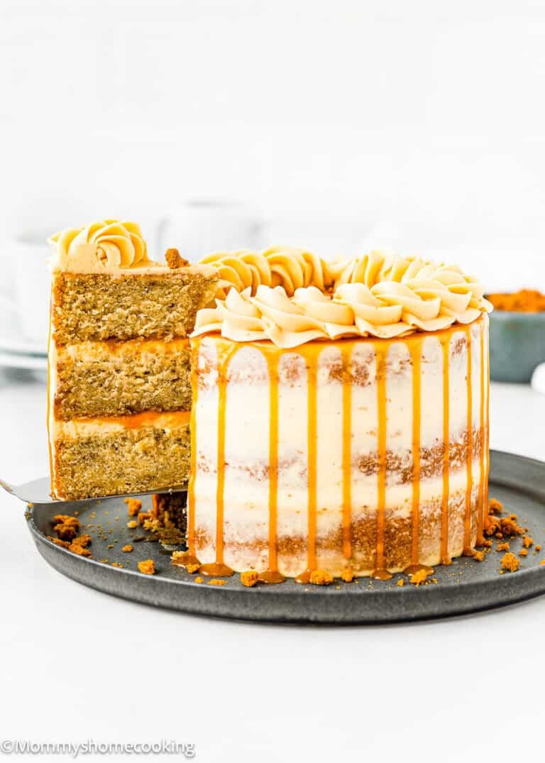 An Eggless Banana Layer Cake with caramel drizzle and piped frosting sits on a dark plate. A slice is being lifted, revealing moist banana layers and creamy filling inside. Blurred cups and a small bowl are in the background.