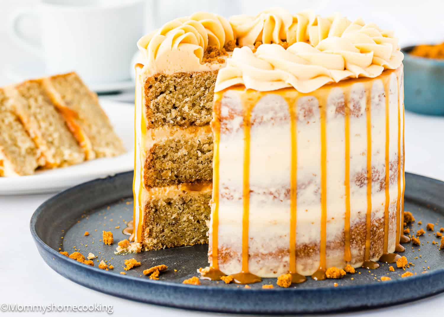 An Eggless Banana Layer Cake with white frosting and caramel drizzle sits on a dark plate. Piped frosting crowns the top, while a slice rests in the background and crumbs are scattered around the plate.