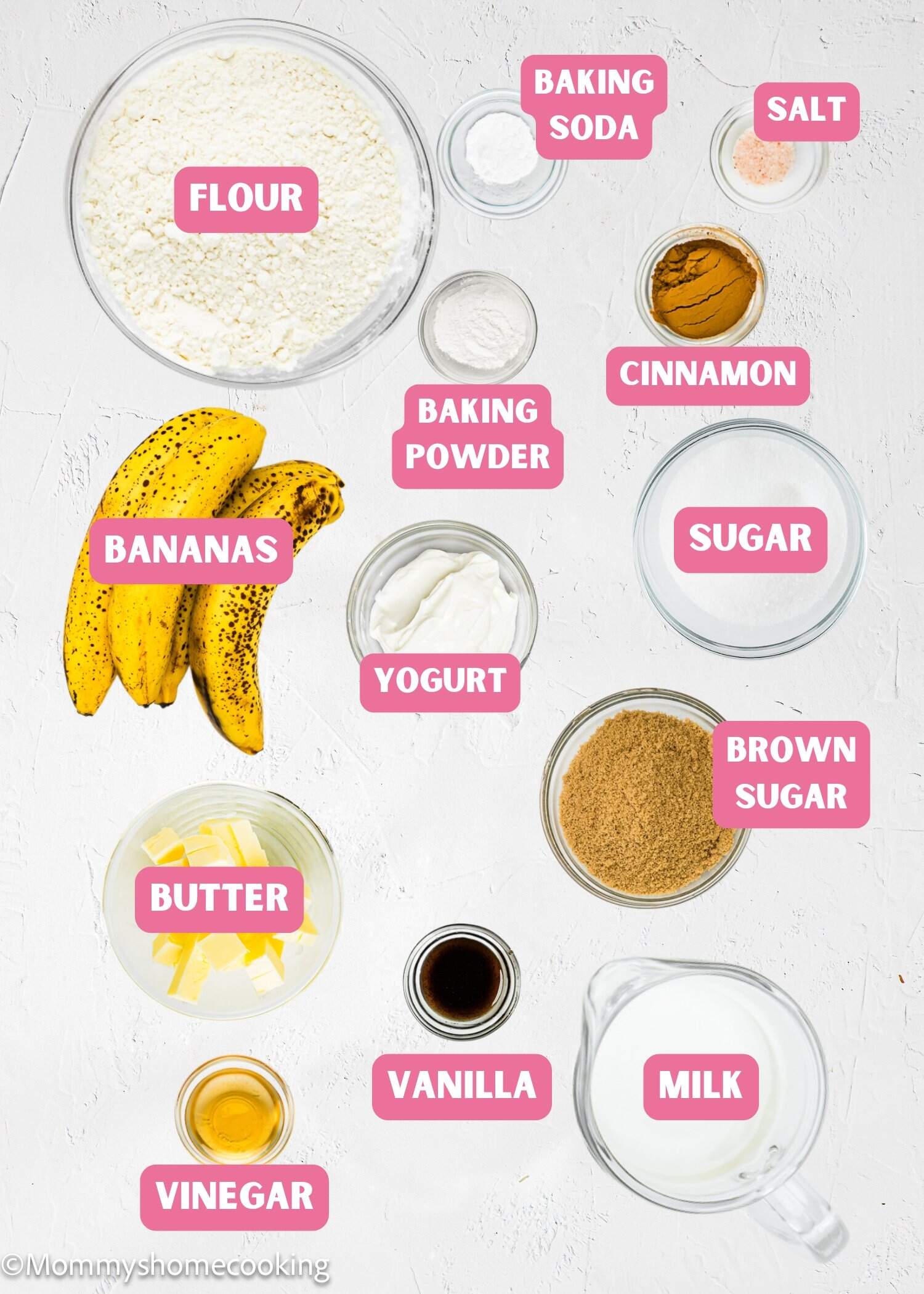 Top-down view of labeled egg-free banana cake ingredients on a white surface, including flour, baking soda, salt, cinnamon, sugar, brown sugar, bananas, yogurt, baking powder, butter, vanilla, milk, and vinegar.
