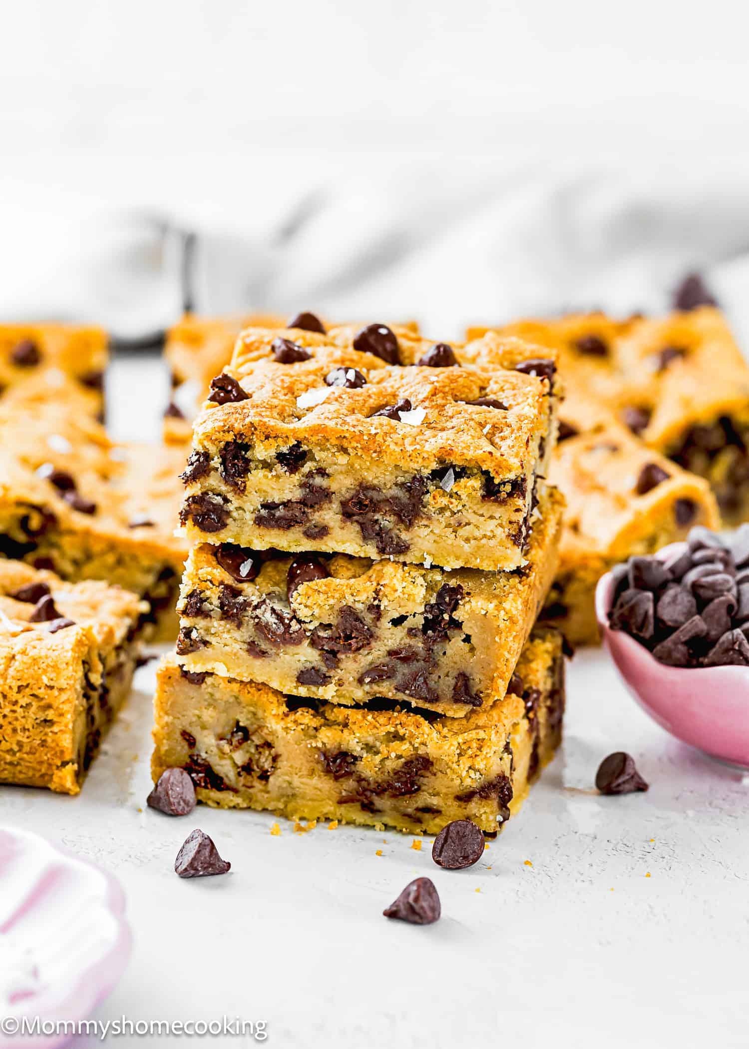A stack of chocolate chip cookie bars with more bars and a small bowl of chocolate chips nearby on a white surface—perfect for anyone searching for Egg-Free Bar Recipes to add to their fall baking lineup.