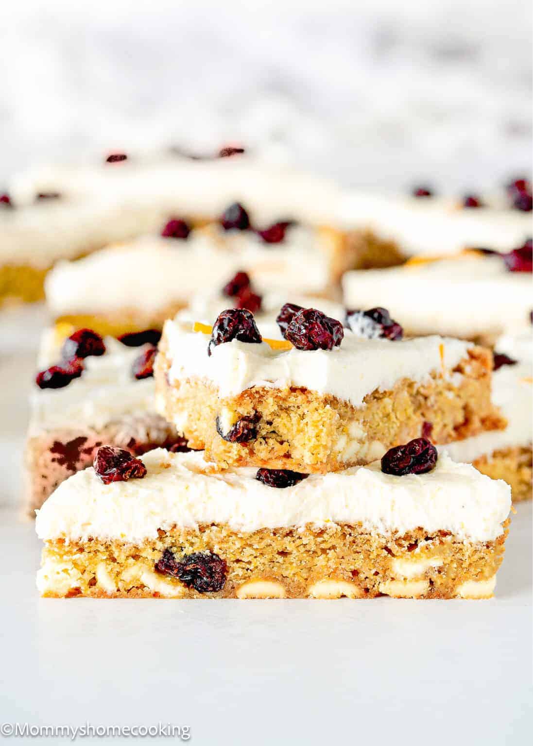 Three dessert bars with white frosting, dried cranberries, and visible white chocolate chips are stacked on a white surface—one of the 10 Easy Egg-Free Bar Recipes Every Baker Needs for a delicious treat.
