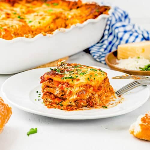 A slice of cheesy egg-free lasagna on a white plate with a fork, garnished with herbs, with the rest of the lasagna in a dish and a block of cheese in the background.