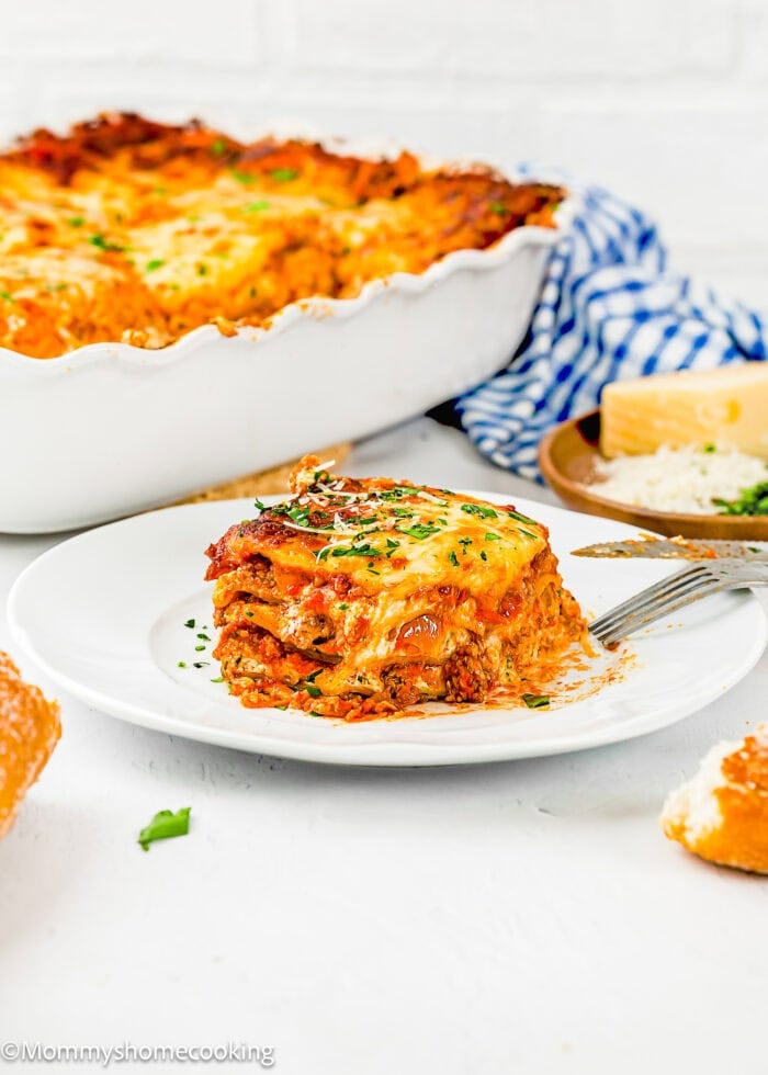 A slice of cheesy egg-free lasagna on a white plate with a fork, garnished with herbs, with the rest of the lasagna in a dish and a block of cheese in the background.