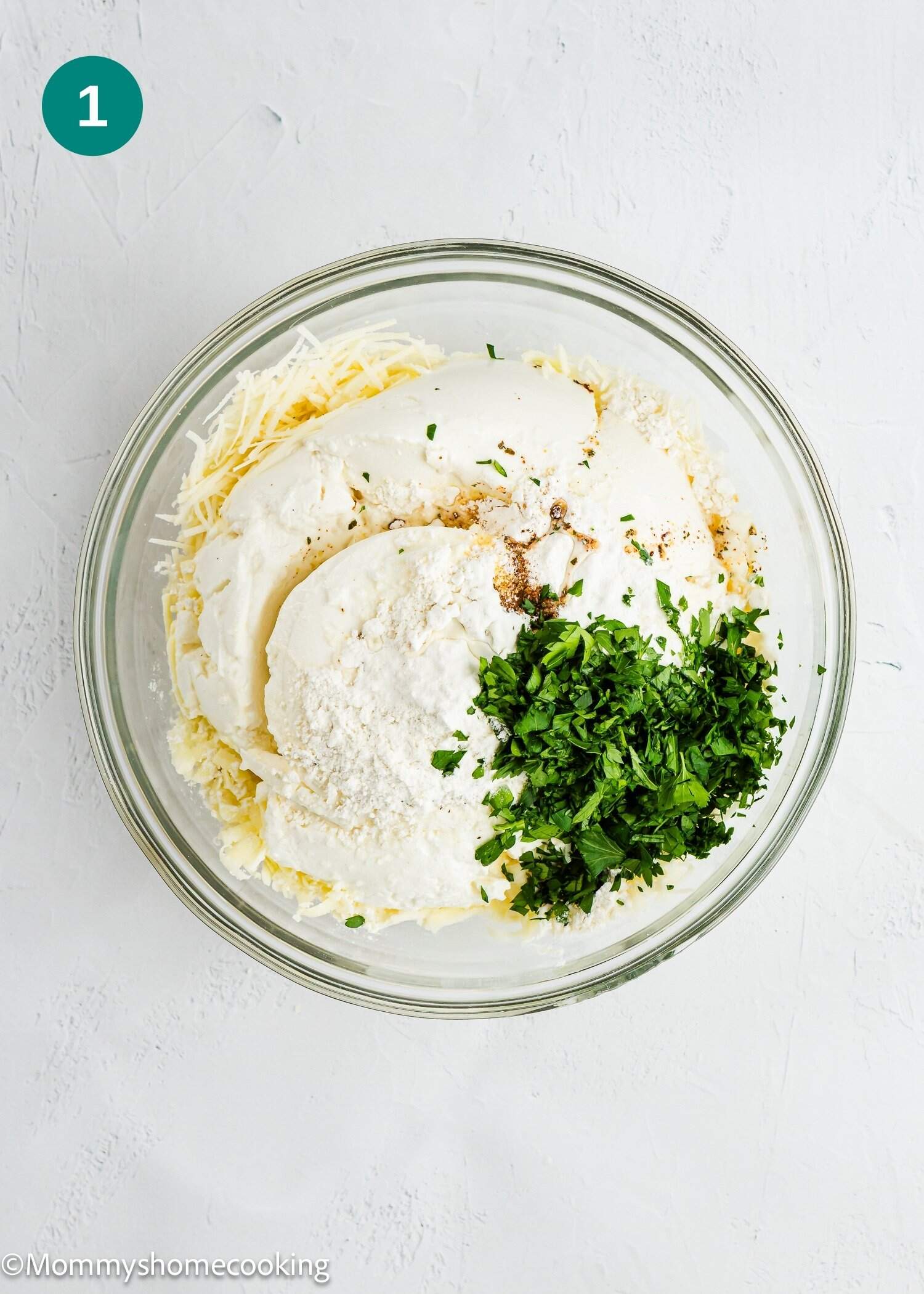 A glass bowl containing ricotta cheese, shredded cheese, flour, chopped parsley, and seasonings on a white surface.