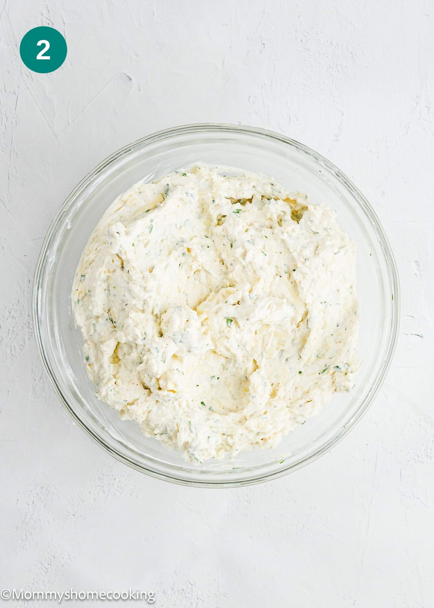 A glass bowl filled with a creamy, mixed cheese and herb spread sits on a white surface.
