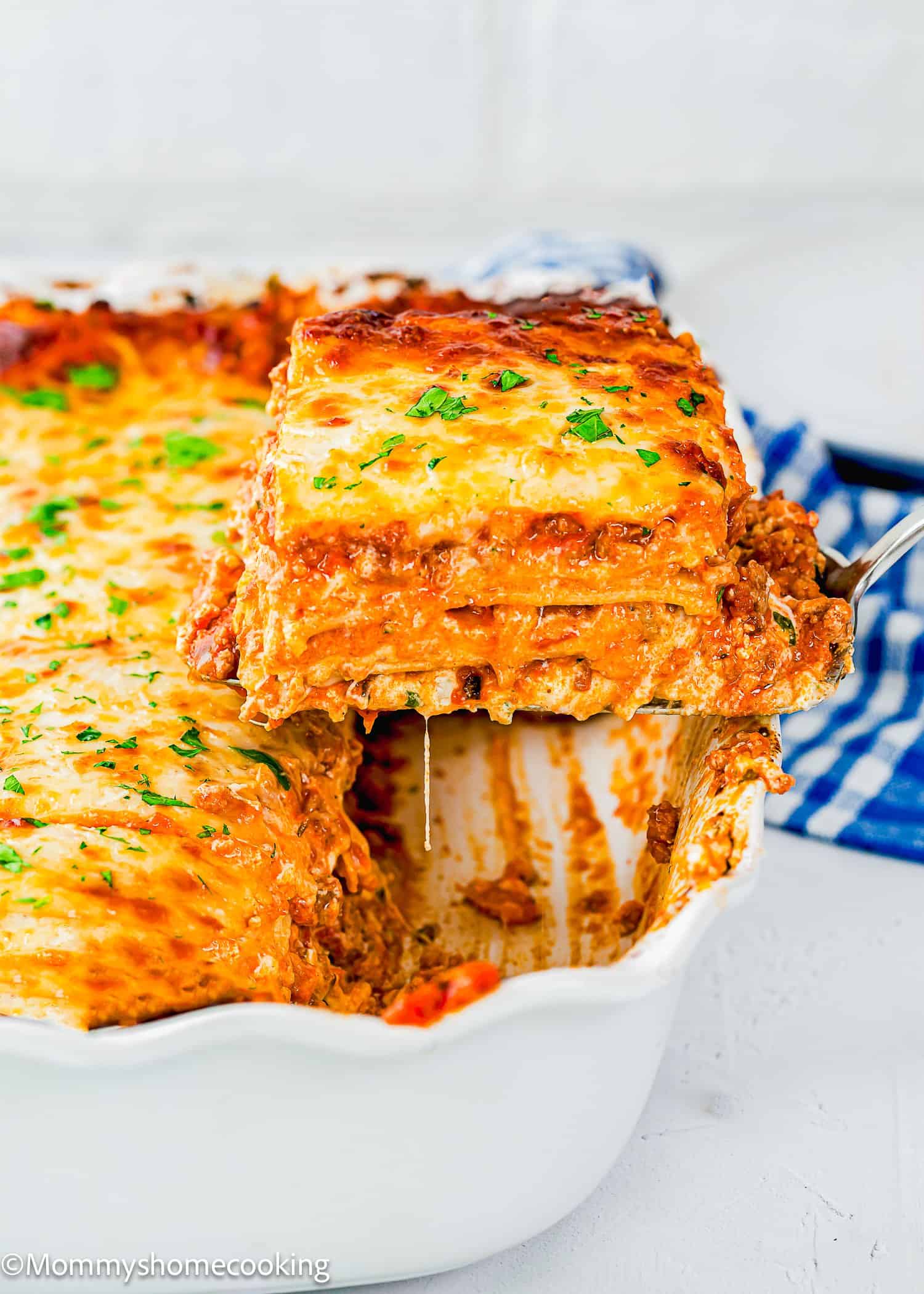 A slice of cheesy, baked egg-free lasagna is being lifted from a white casserole dish, showing layers of pasta, meat sauce, and melted cheese.