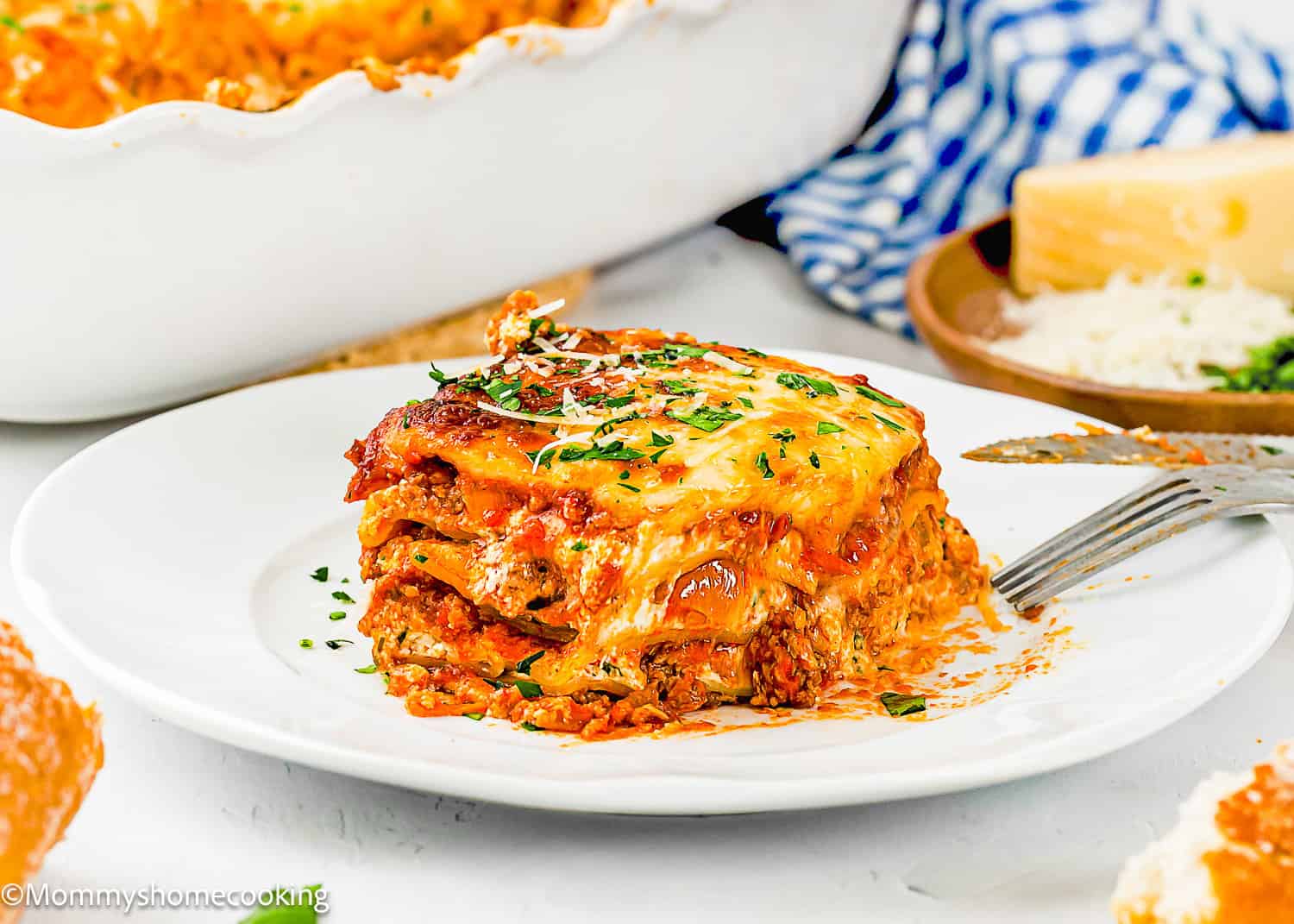 A slice of eggless lasagna with layers of meat, cheese, and sauce sits on a white plate, garnished with chopped herbs; a fork and cheese are nearby.