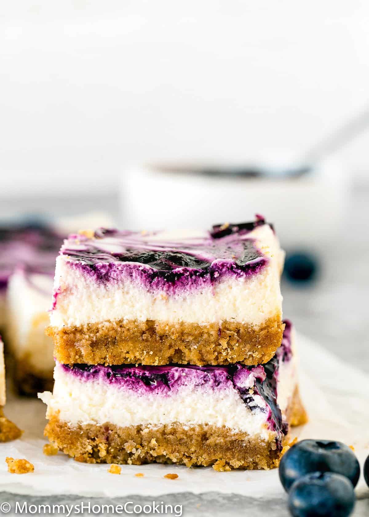 Two blueberry swirl cheesecake bars with a graham cracker crust are stacked on top of each other, next to fresh blueberries—a tempting treat from our 10 Egg-Free Bar Recipes That Are Simply Irresistible.