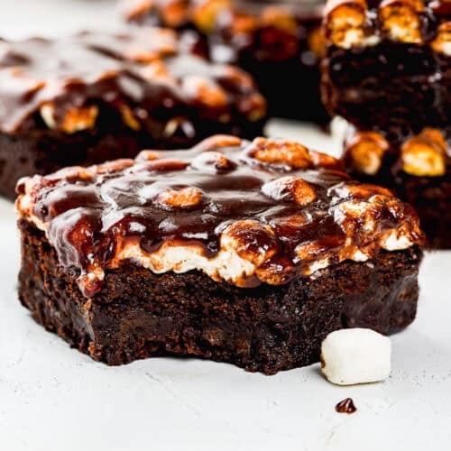 A egg-free chocolate brownie topped with toasted marshmallows and chocolate glaze, with a bite taken out, sitting on a white surface.