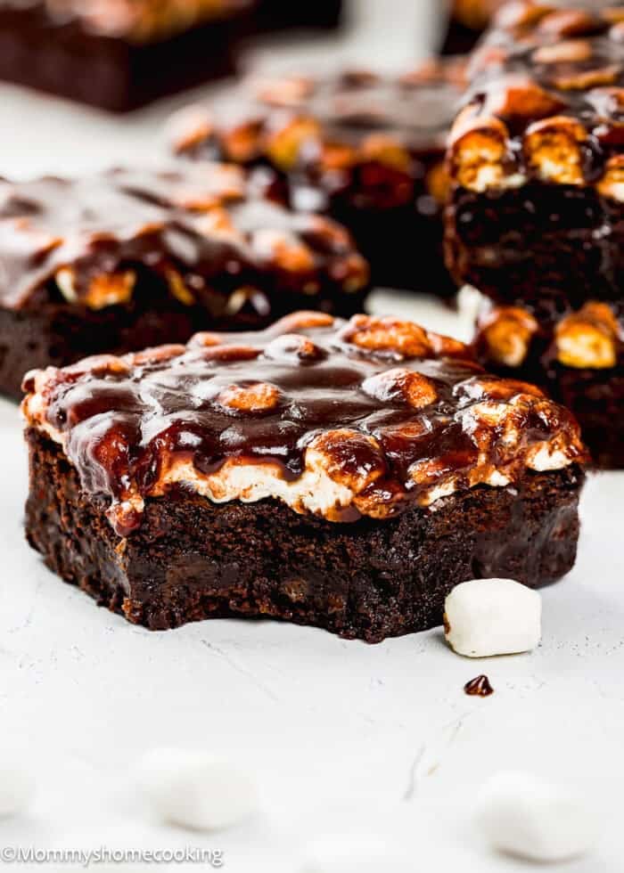 A egg-free chocolate brownie topped with toasted marshmallows and chocolate glaze, with a bite taken out, sitting on a white surface.