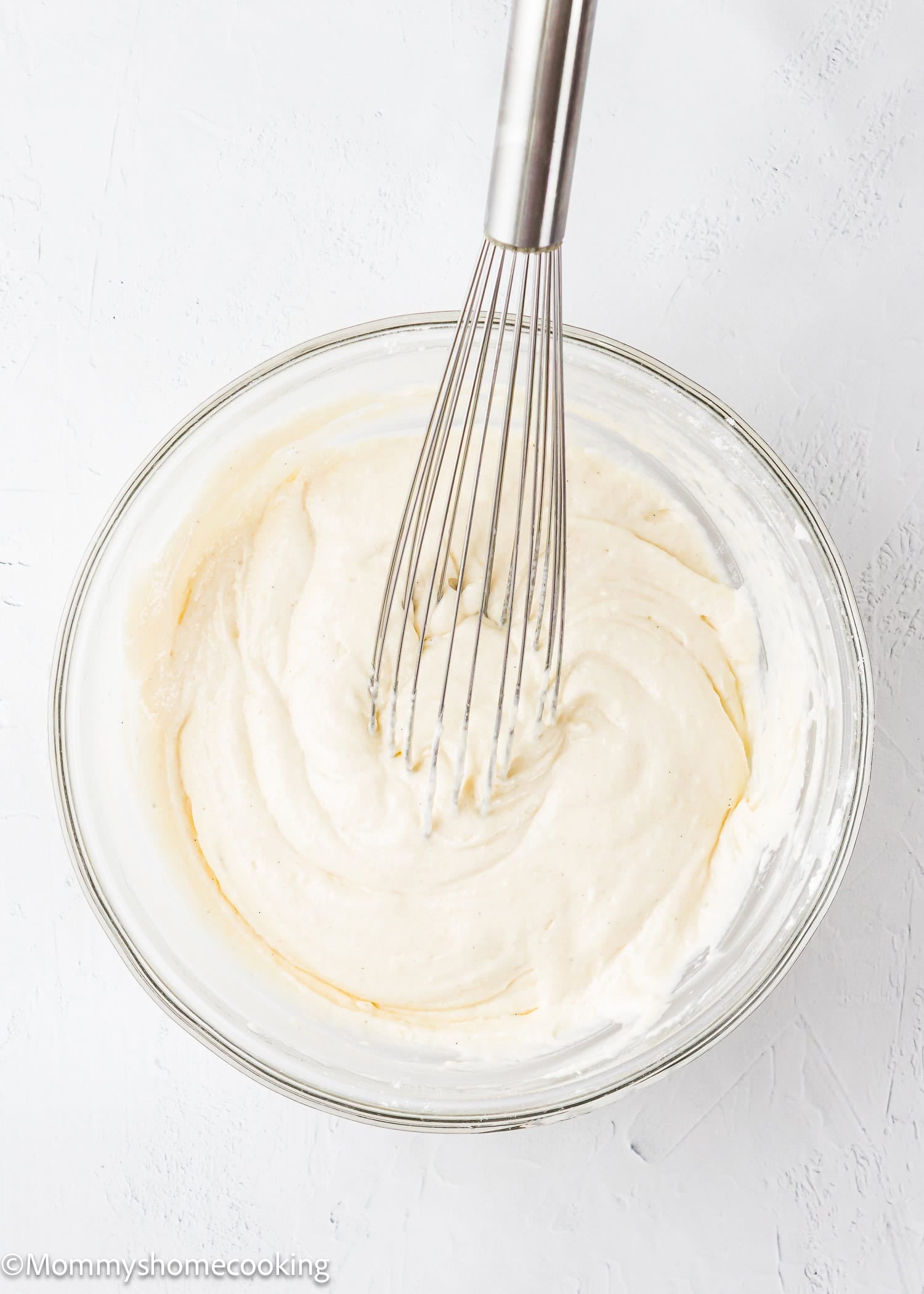 A metal whisk in a glass bowl filled with creamy white batter for Eggless Protein Waffles rests on a light surface.