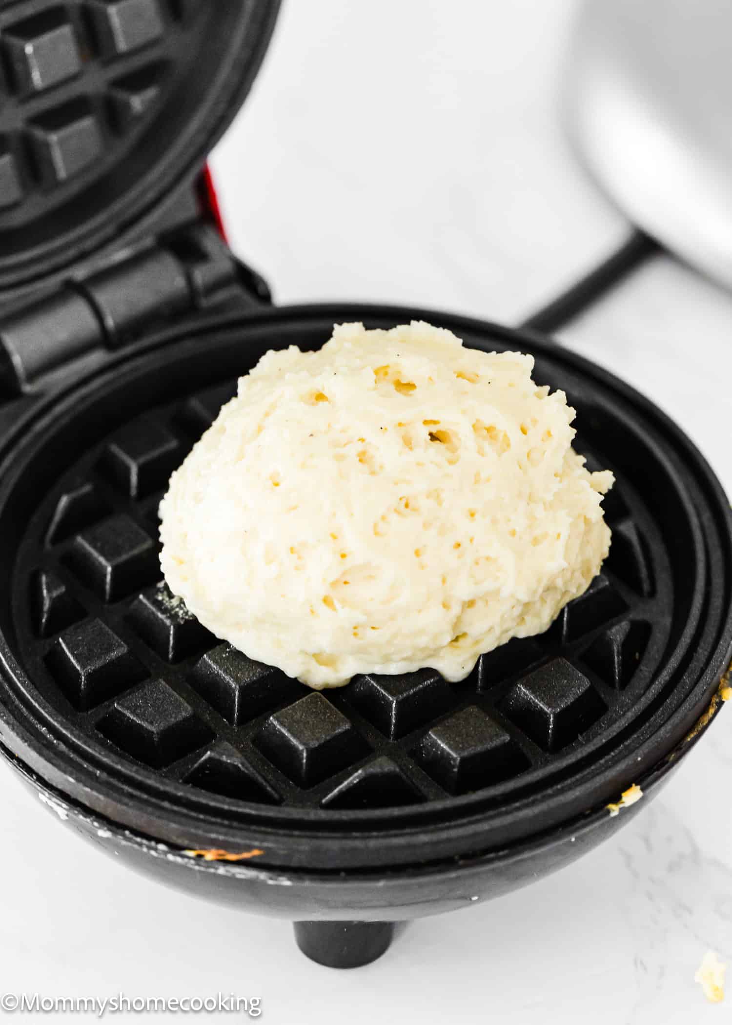 A dollop of Eggless Protein Waffles batter sits on the grid of an open, round waffle maker, ready to be cooked. The black waffle maker gleams against a light-colored countertop, promising a healthy twist on breakfast.