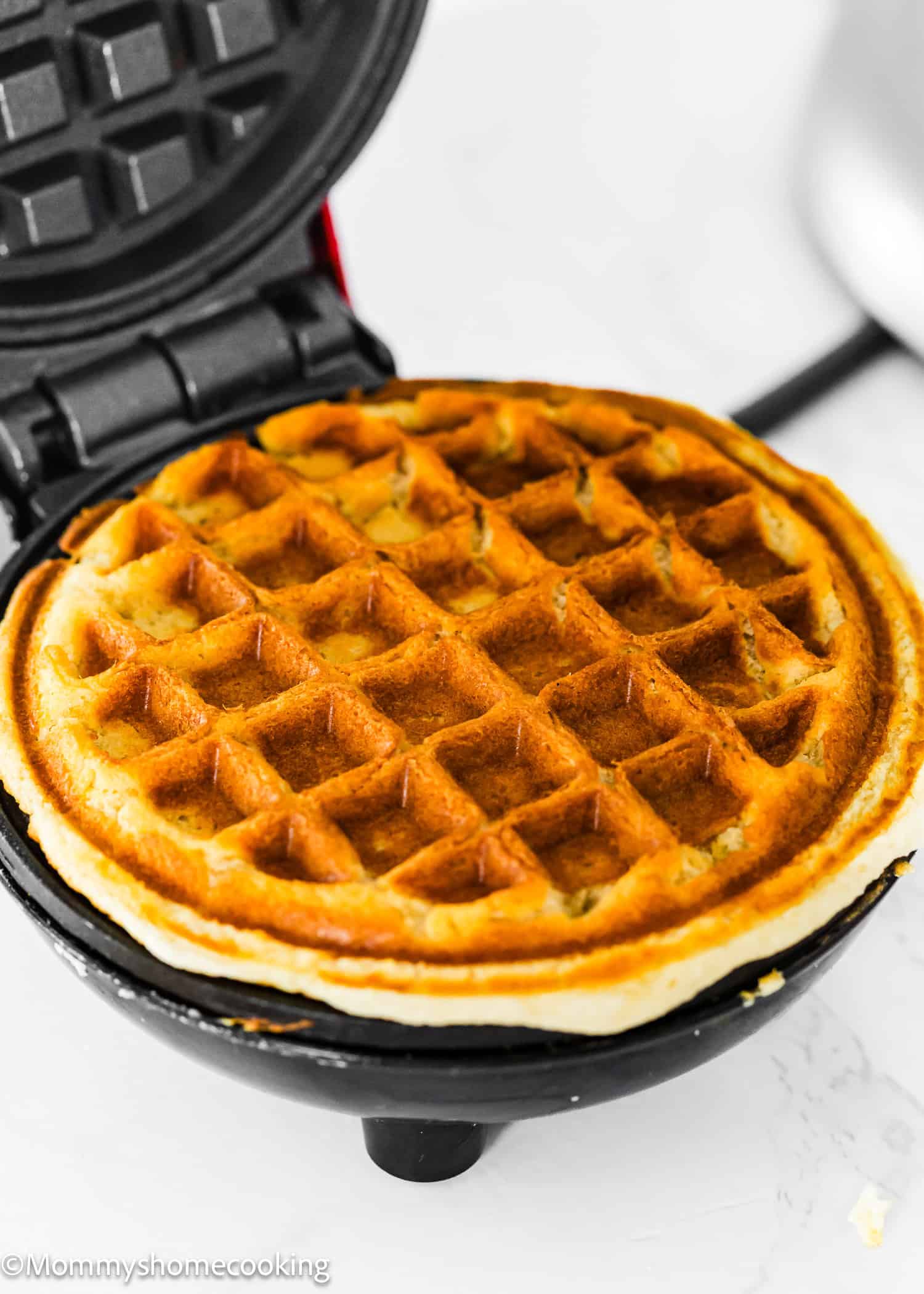 A golden-brown Eggless Protein Waffle cooking in an open waffle maker, ready to be served. The background is a clean, white surface.