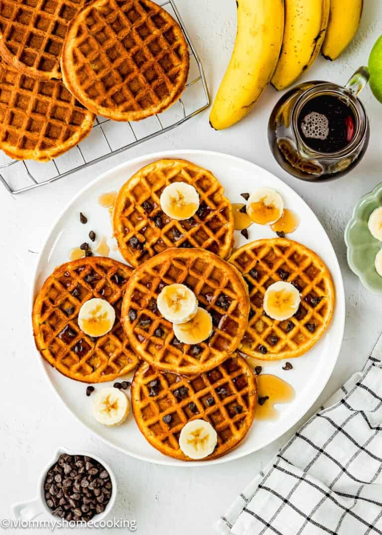 A plate of six Egg-Free Protein Waffles topped with banana slices and syrup, surrounded by chocolate chips, bananas, a small jug of syrup, and a checkered napkin on a white surface.