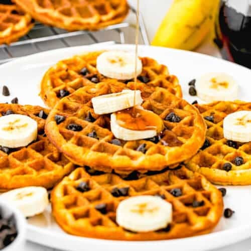 A plate of golden Egg-Free Protein Waffles topped with banana slices and chocolate chips is drizzled with syrup. A banana and a jar of syrup are in the background. The scene is bright and inviting.