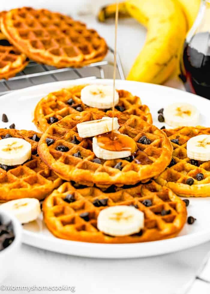 A plate of golden Egg-Free Protein Waffles topped with banana slices and chocolate chips is drizzled with syrup. A banana and a jar of syrup are in the background. The scene is bright and inviting.