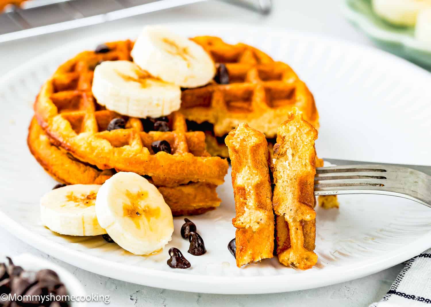 A plate with two Egg-Free Protein Waffles topped with banana slices and chocolate chips. A fork holds a bite-sized piece of waffle, with more banana slices and chocolate chips scattered on the plate.