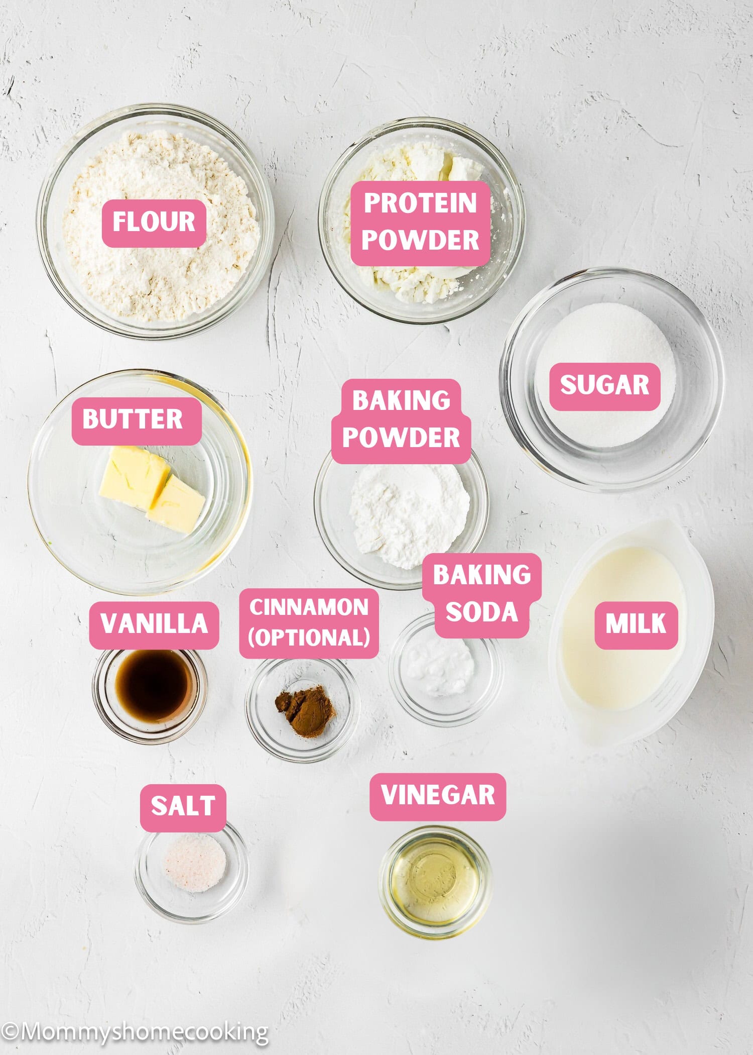 Top-down view of labeled baking ingredients in glass bowls on a white surface, perfect for making Eggless Protein Waffles—featuring flour, protein powder, sugar, butter, baking powder, soda, milk, vanilla, cinnamon, salt, and vinegar.