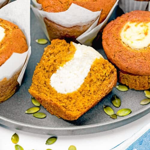 Egg-Free Pumpkin muffins with cream cheese filling on a round tray. One muffin is cut in half, showing the creamy center. Pumpkin seeds are scattered beside the muffins.