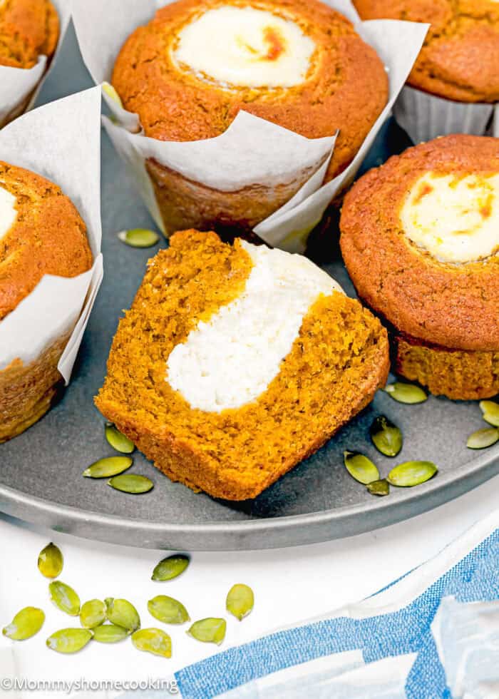 Egg-Free Pumpkin muffins with cream cheese filling on a round tray. One muffin is cut in half, showing the creamy center. Pumpkin seeds are scattered beside the muffins.