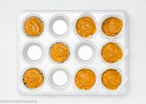 A muffin tin with 12 cups, 6 of which are filled with orange batter in paper liners, while the other 6 cups are empty. The tin is on a white surface.