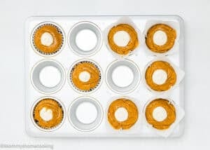 A muffin tin with eight unbaked pumpkin muffins, each topped with a dollop of white cream cheese mixture. Four muffin spaces are empty. The tin is on a white background.