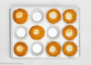 A muffin tin containing seven pumpkin muffins, three of which have a cream cheese center, with three empty muffin cups. The tin is placed on a cooling rack over a white background.