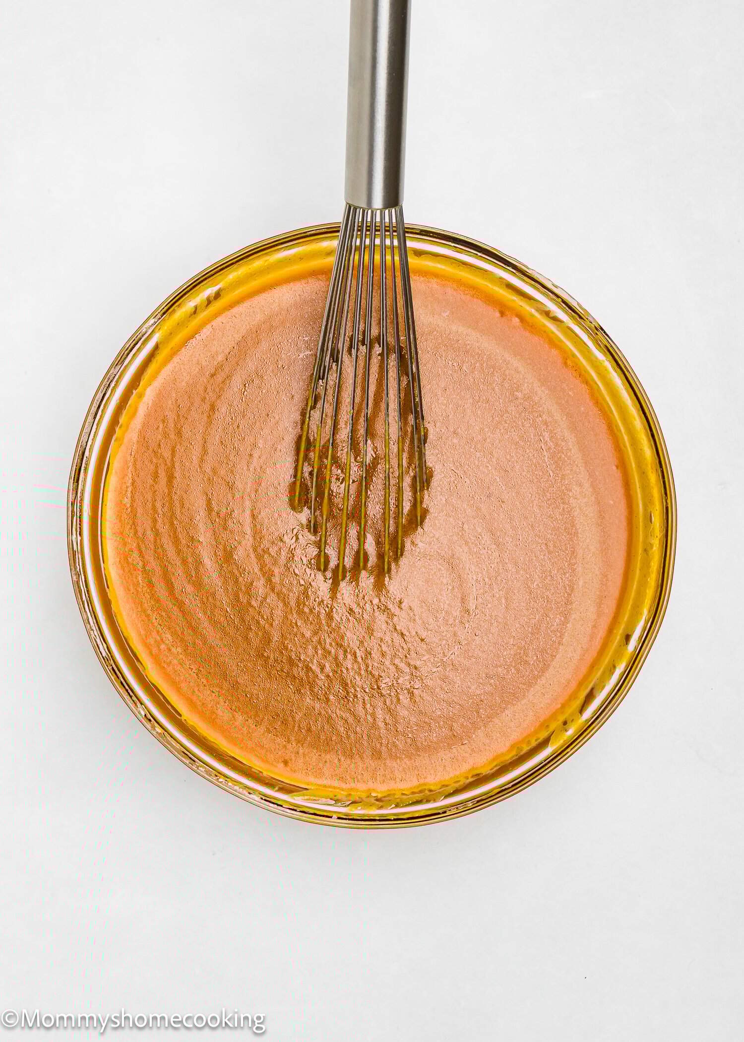 A metal whisk rests in a glass bowl filled with smooth, light brown cake batter on a white surface.