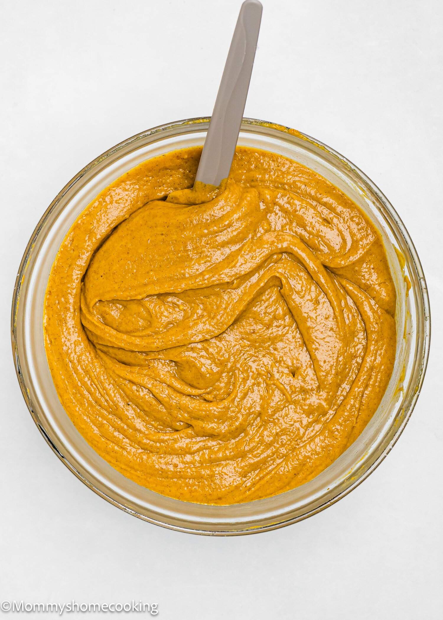 A glass bowl filled with smooth pumpkin batter, with a spatula resting inside, on a white surface.