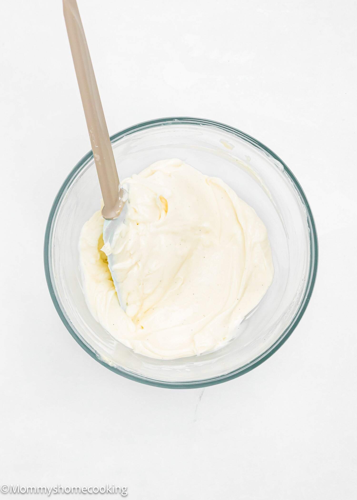 A glass bowl filled with creamy white mixture, being stirred with a beige spatula, on a white background.