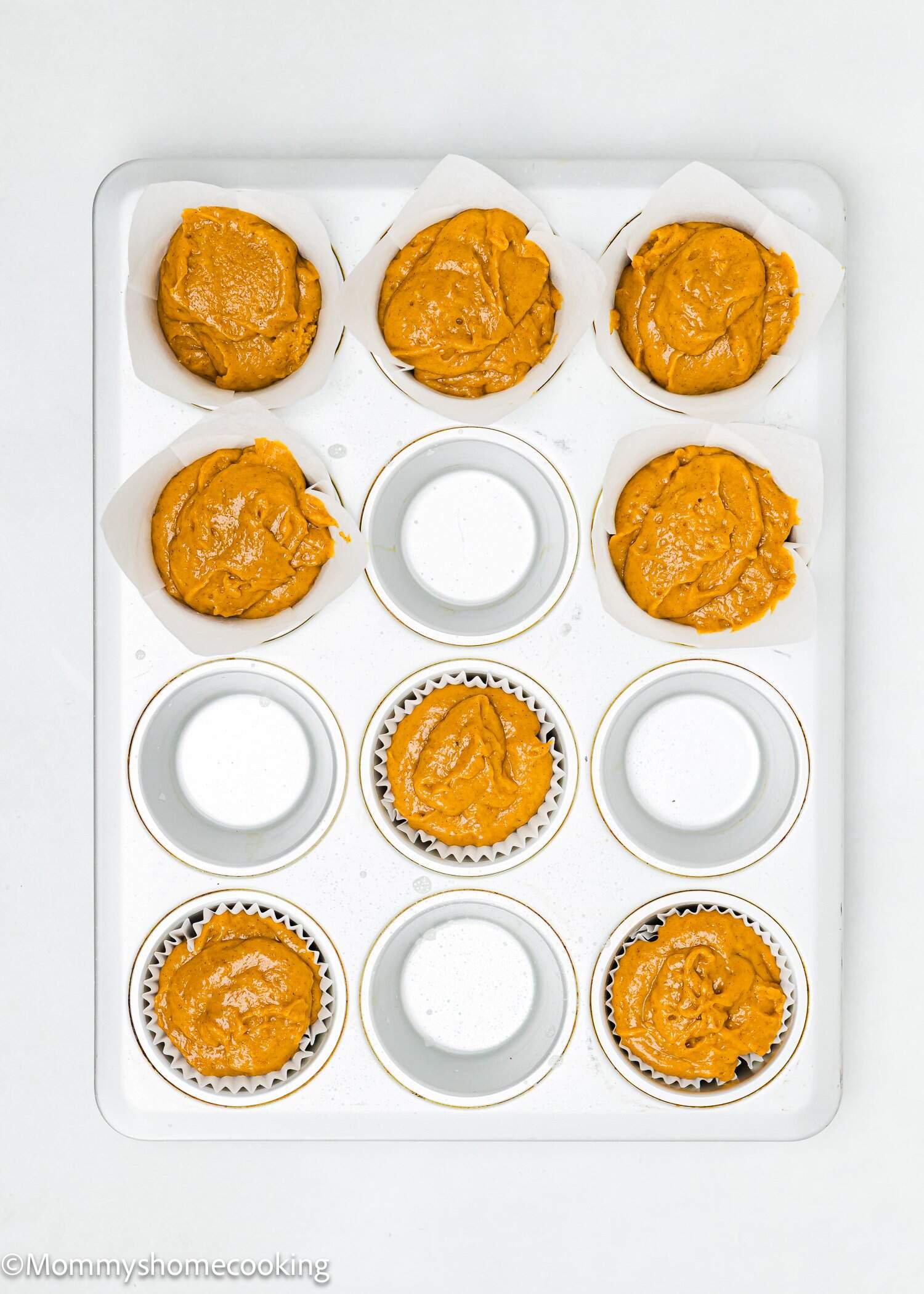 A muffin tin with six cups filled with orange batter in white paper liners, and six empty cups. The tin is placed on a white surface.