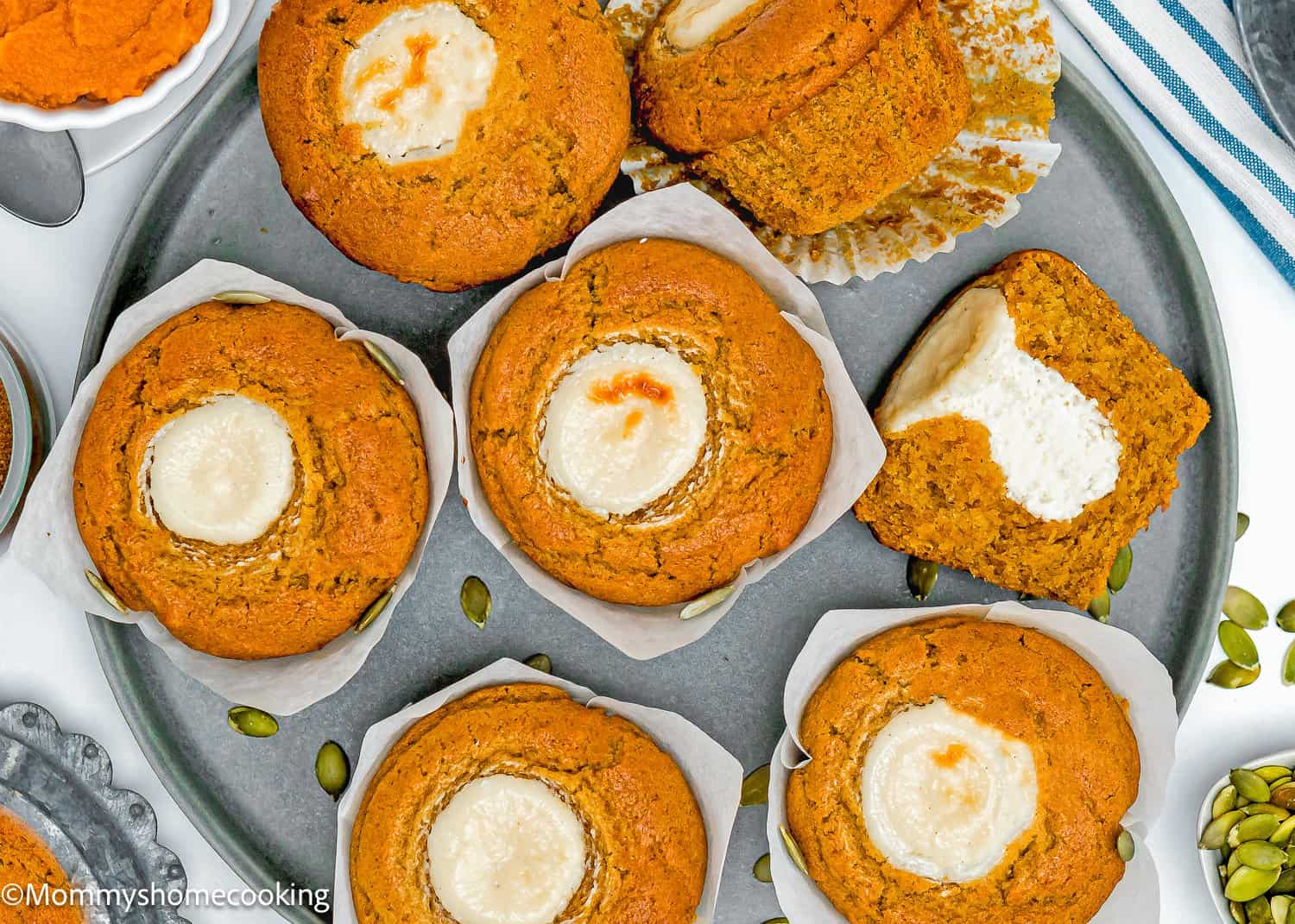 Eggless Pumpkin muffins with cream cheese centers arranged on a round metal tray; one muffin is cut in half to show the filling. Pumpkin seeds are scattered nearby.