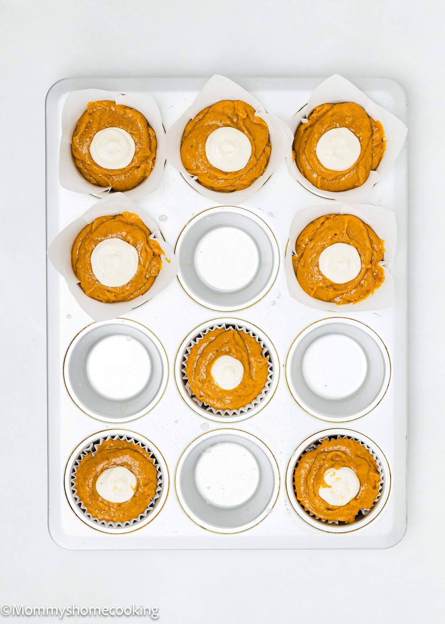 A muffin tin with seven cups filled with pumpkin muffin batter, each topped with a dollop of white cream cheese mixture. Five cups are empty. The tin is on a white background.