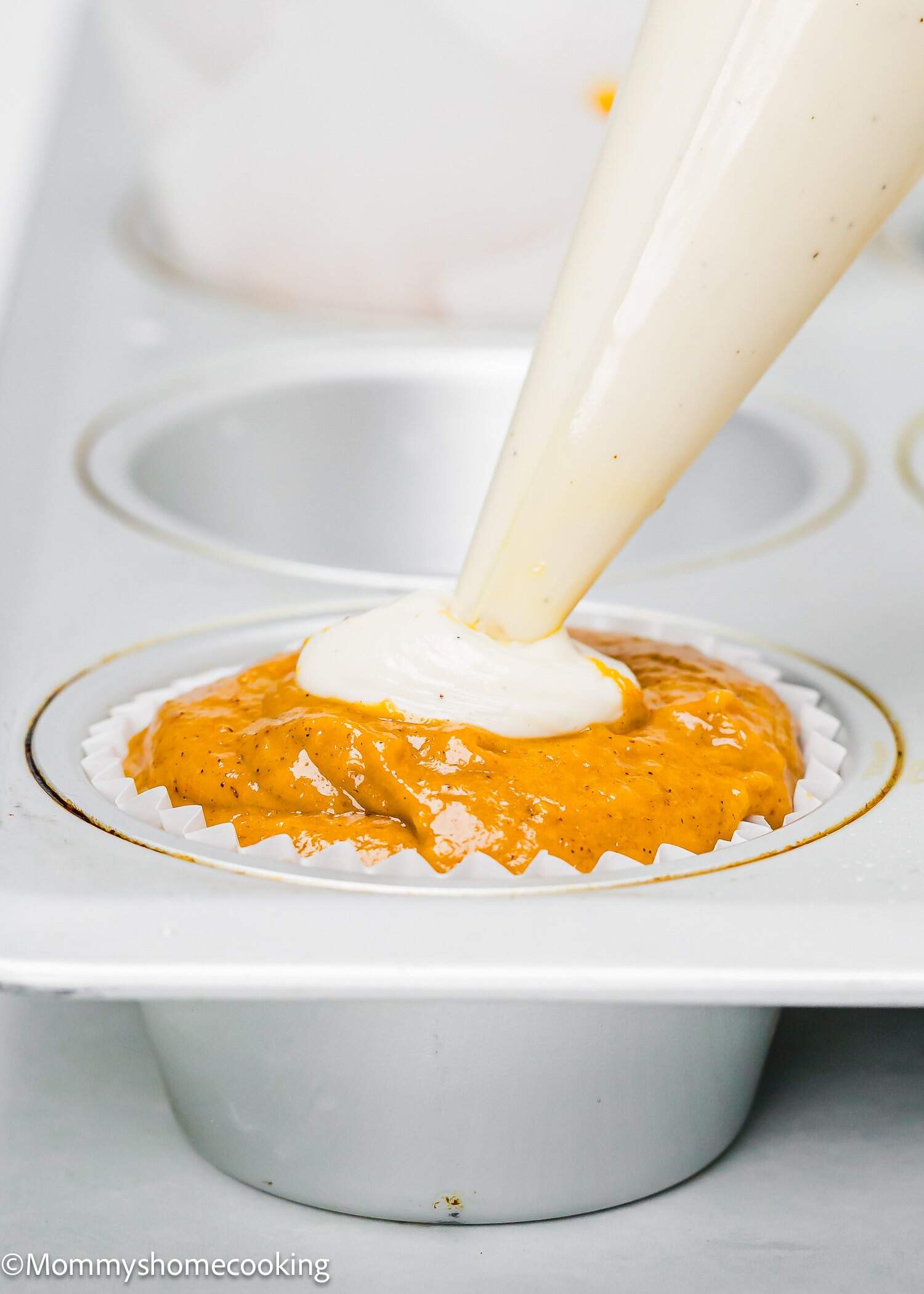 A piping bag fills the center of pumpkin muffin batter with cream cheese in a white paper liner inside a muffin tin.
