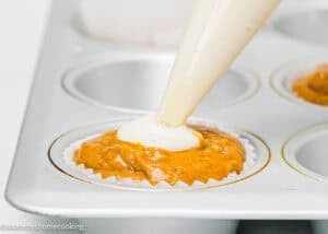 A piping bag is filling the center of pumpkin muffin batter with cream cheese in a cupcake liner inside a muffin tray.