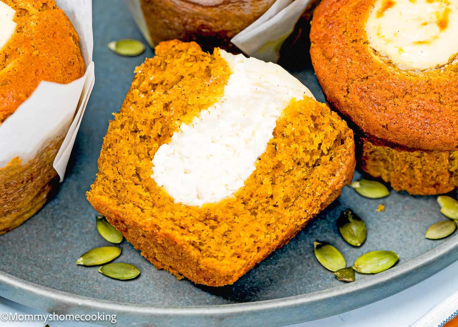 A sliced egg-free pumpkin muffin with a creamy white filling is displayed on a blue plate, surrounded by pumpkin seeds and other muffins.