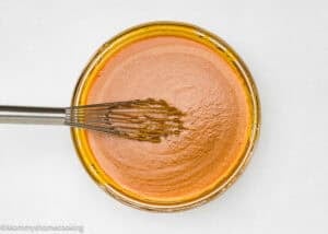 A metal whisk resting in a glass bowl filled with smooth, light brown batter, viewed from above on a white surface.