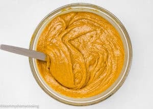 A glass bowl filled with smooth, orange-brown batter being mixed with a spatula, set on a white surface.